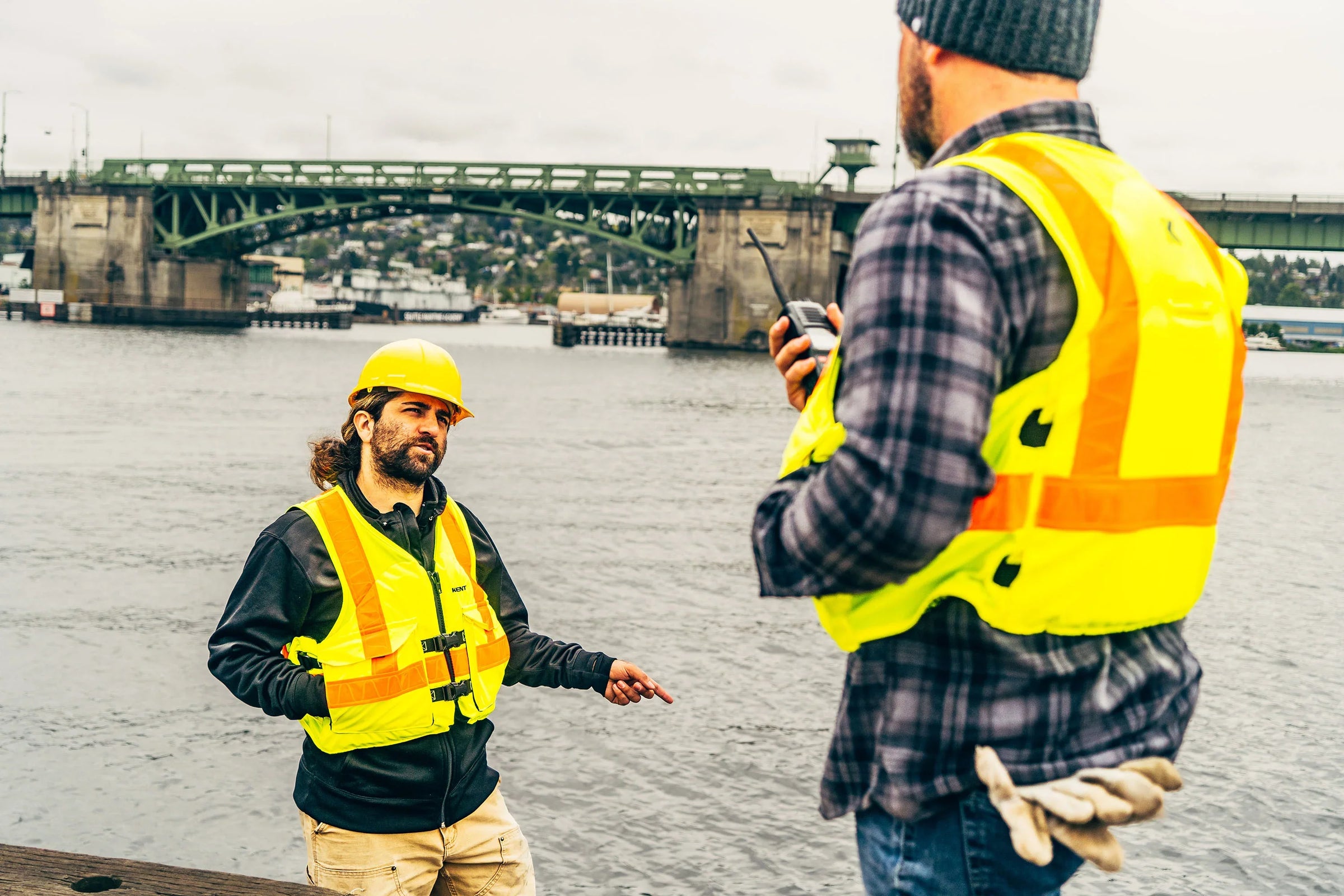 Two workers on a bridge above water, wearing Yellow and Orange Reflective Life Vests.